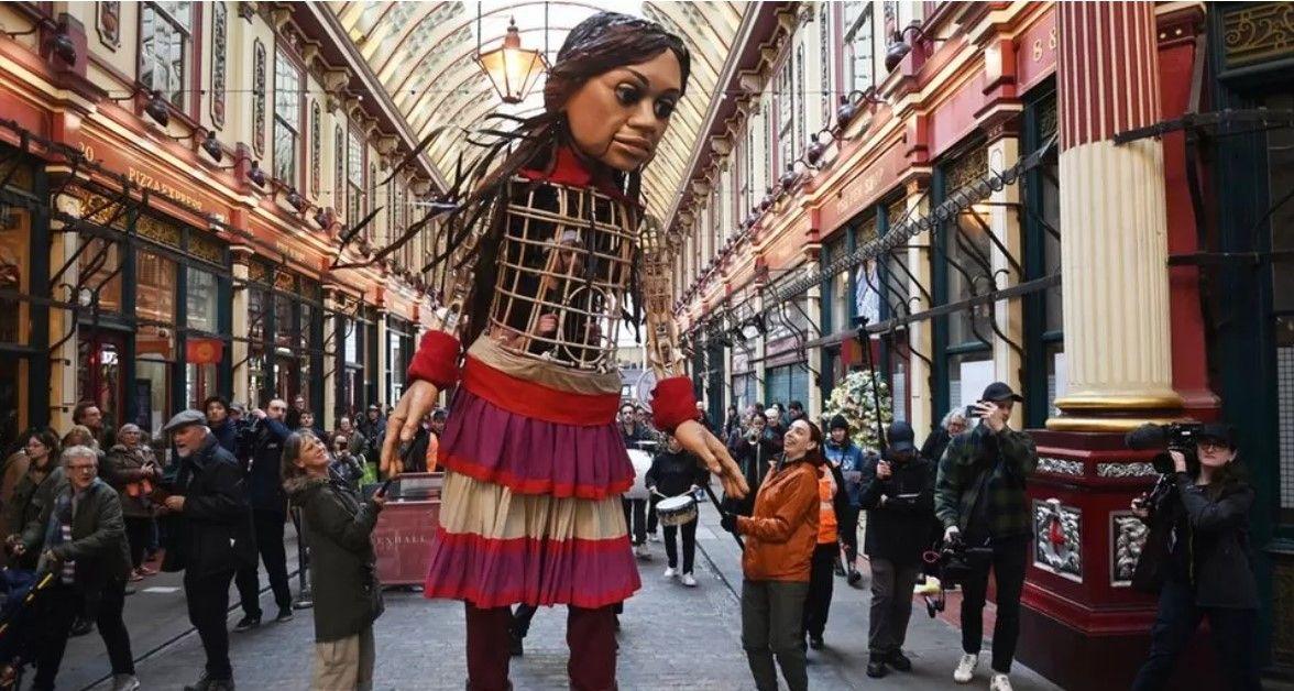 A 12ft (3.7m) tall puppet of a girl stands in an old shopping centre in London as people look on