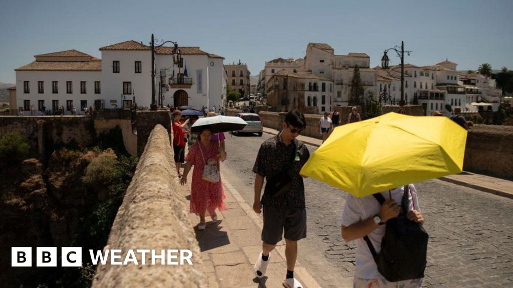 People hide from the sun with umbrellas as they cross an old bridge. White buildings in the background