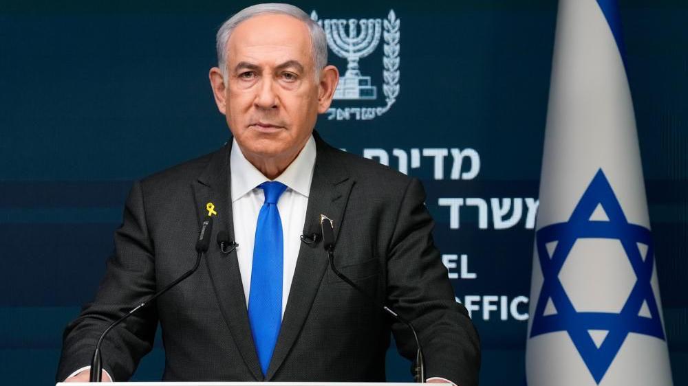 Israeli Prime Minister Benjamin Netanyahu speaking from a podium during a press conference. He is wearing a black suit and blue tie. Behind him is an Israeli flag.