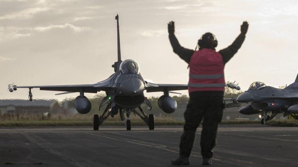 A sleek military jet taxis on the RAF Waddington runway for Exercise Cobra Warrior. A member of the ground crew, wearing a pink hi-vis vest, holds his hands in the air as he signals to the pilot.