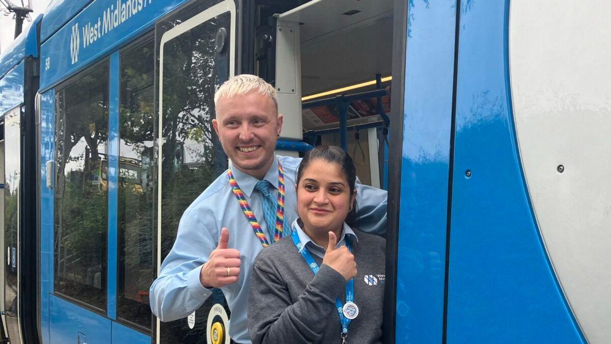 A man with bleach blond hair standing behind a woman with black hair. They are both hanging out the side of a blue tram while smiling and sticking their thumbs up.