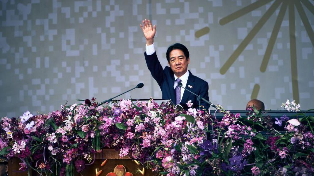 William Lai, in a suit, waves an arm towards the crowd while on stage