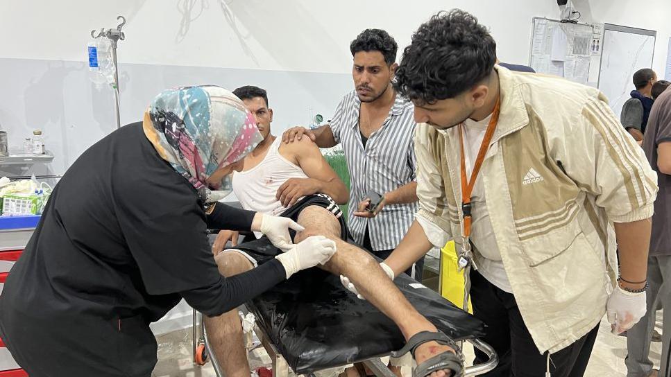 An injured man is lying on a hospital trolley while medics treat his wounds, another man is looking on concerned