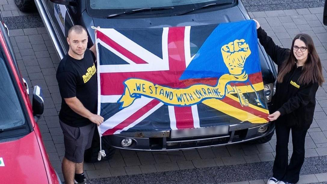 Kayleigh Jones (right) and Joshua Katihrob standing wearing black clothes with a large flag in front of a black truck. Flag is a Union Jack with the words "we stand with Ukraine" written on it