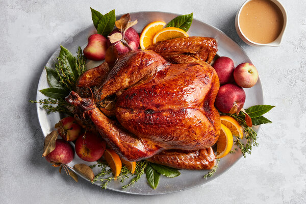 An overhead image of a turkey on a platter surrounded by herbs, apples and oranges.