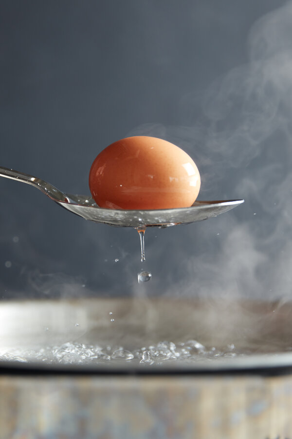 A whole brown egg on a spoon emerges from a boiling pot of water. Steam rises from the pot in the background.